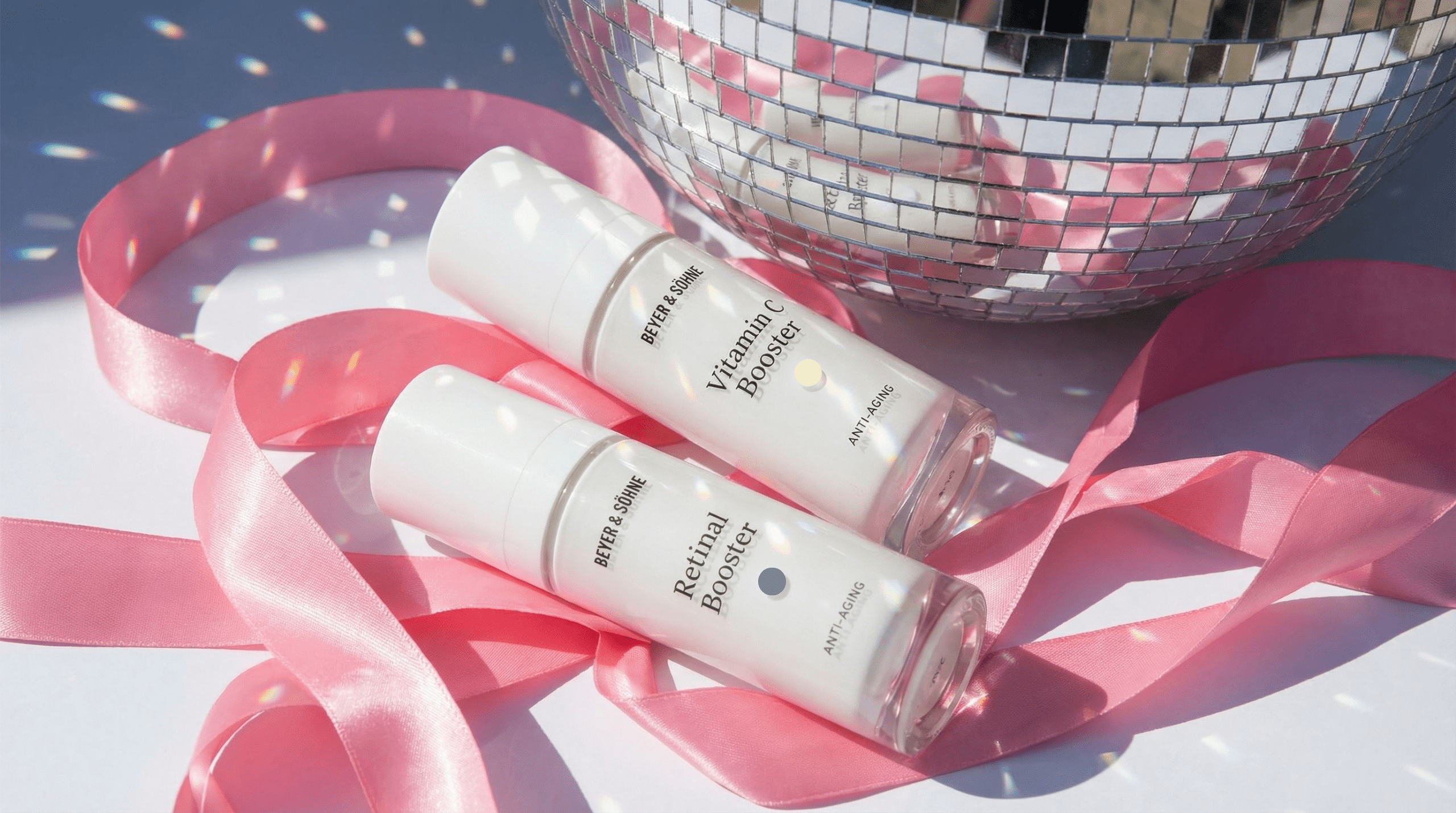 Beyer & Söhne Vitamin C und Retinal Booster. Anti-Aging Seren in festlicher Szene mit Discokugel und rosa Geschenkband.